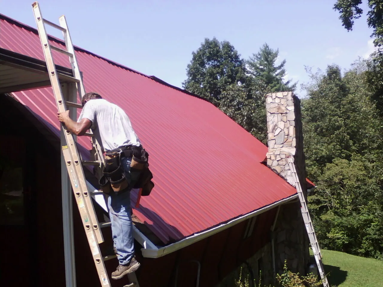 Roofer on a ladder beside a red metal roof and stone chimney — Soffit & Fascia Repair by Roofing Brothers in Ricketts
